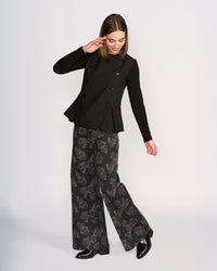 A woman smiles and tilts her head, raising one arm as she models the Knewe Label Austin Top in black against a plain background, paired with wide-leg black pants featuring a subtle floral pattern.