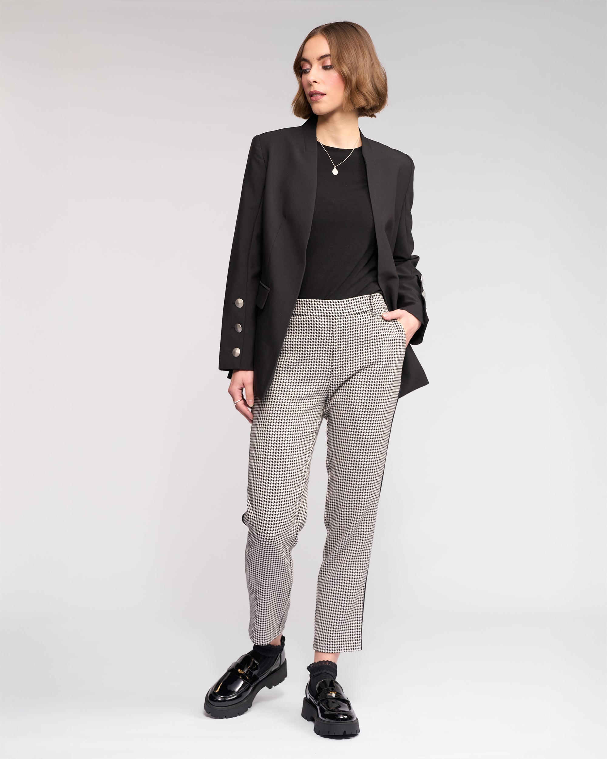 Against a plain background, a person wears the Carter Blazer - Black by Knewe Label, paired with a black top, checked pants, and chunky black shoes. With one hand in their pocket, they look to the side in a relaxed pose.