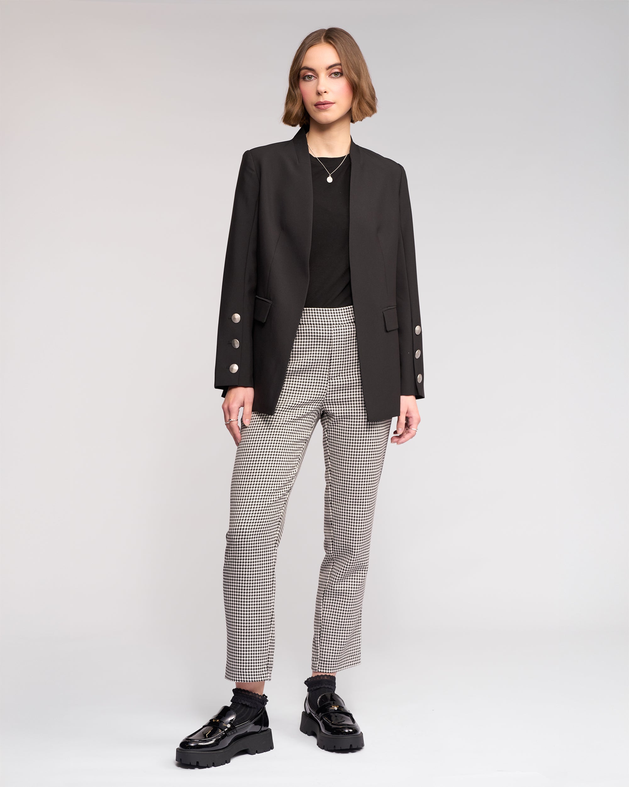 A woman with short hair and a neutral expression models the Knewe Label Carter Blazer in black, featuring silver buttons, over a black top and checkered pants with black chunky shoes, accentuating her modern silhouette against a plain background.