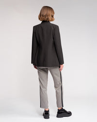 A person with short light brown hair wears the Knewe Label Carter Blazer in black, paired with patterned side-stripe pants and black platform shoes, showcasing a modern silhouette against a plain light background.