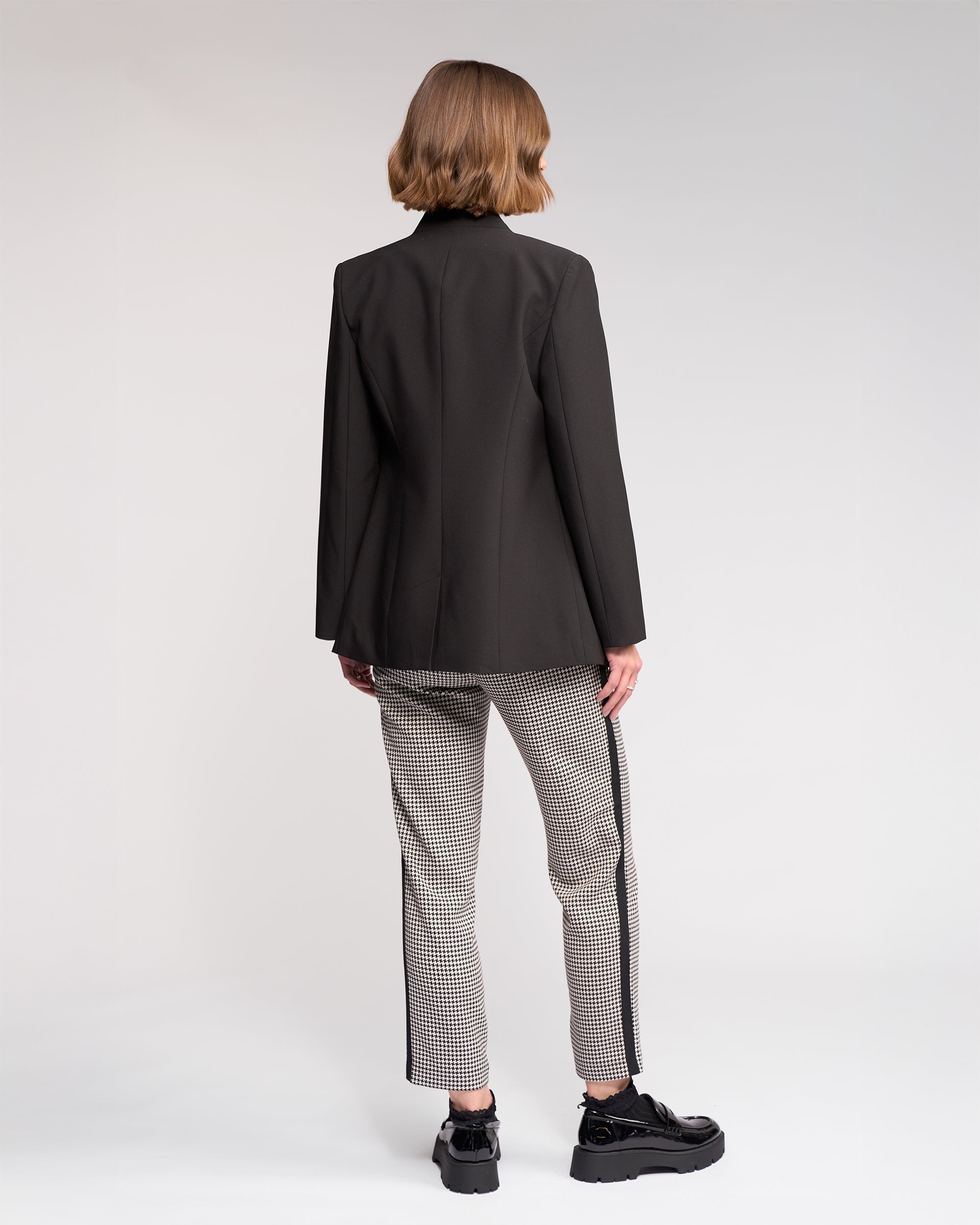 A person with short light brown hair wears the Knewe Label Carter Blazer in black, paired with patterned side-stripe pants and black platform shoes, showcasing a modern silhouette against a plain light background.