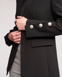A person wearing the Knewe Label Carter Blazer in black, which is collarless with three large silver sleeve buttons, paired with patterned pants and silver rings, shown from shoulders to mid-thigh against a neutral background.