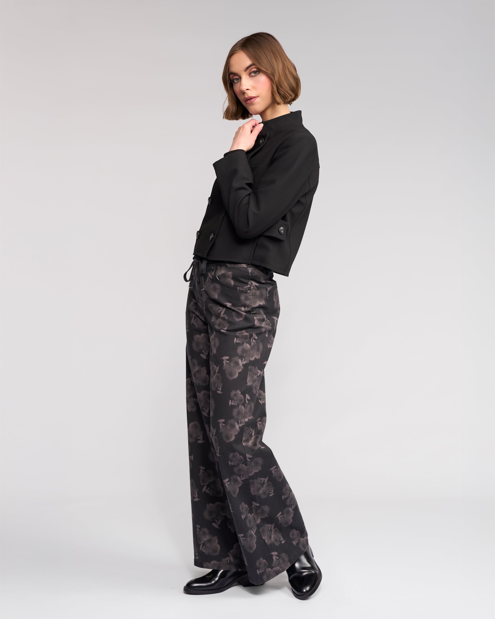 A woman with short brown hair models the Knewe Label Soho Jacket in black, posing in a studio with one hand near her chin against a plain light gray background. She pairs the jacket with wide-leg patterned pants and black shoes.