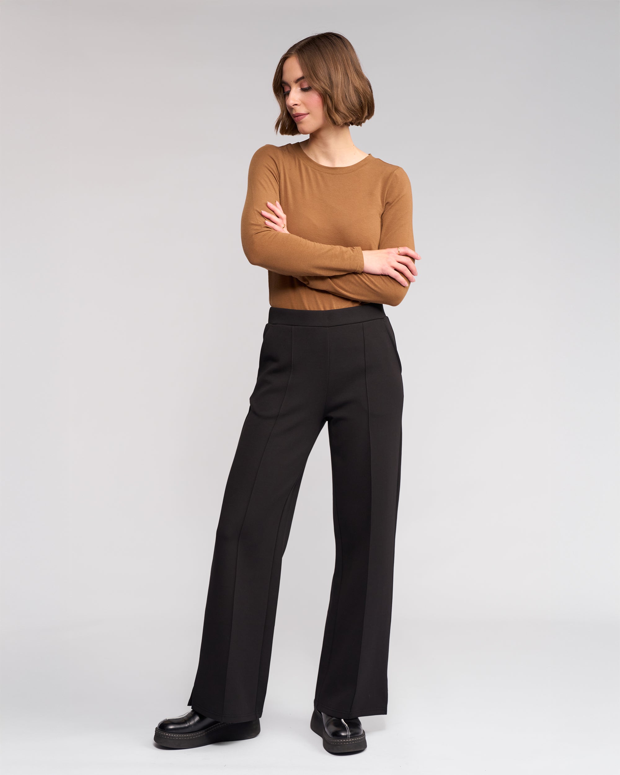A person with chin-length brown hair stands with arms crossed, wearing a fitted brown long-sleeve top, Knewe Label Dune Pant - Black, and black platform shoes against a plain light gray background.