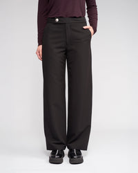 Someone wearing the Knewe Label Blaze Pant in Black stands with one hand in their pocket, paired with a dark long-sleeve top and black shoes, against a plain light background.