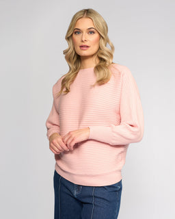 A woman with long blonde hair wears the Vassalli Kiki Sweater in dusky pink and blue jeans, standing against a plain light gray background. She faces forward with a neutral expression and one hand on her hip.