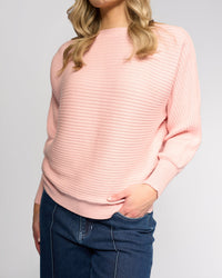 A woman stands against a plain background wearing the Vassalli Kiki Sweater in dusky pink with blue jeans, her face partially visible and one hand in her pocket.