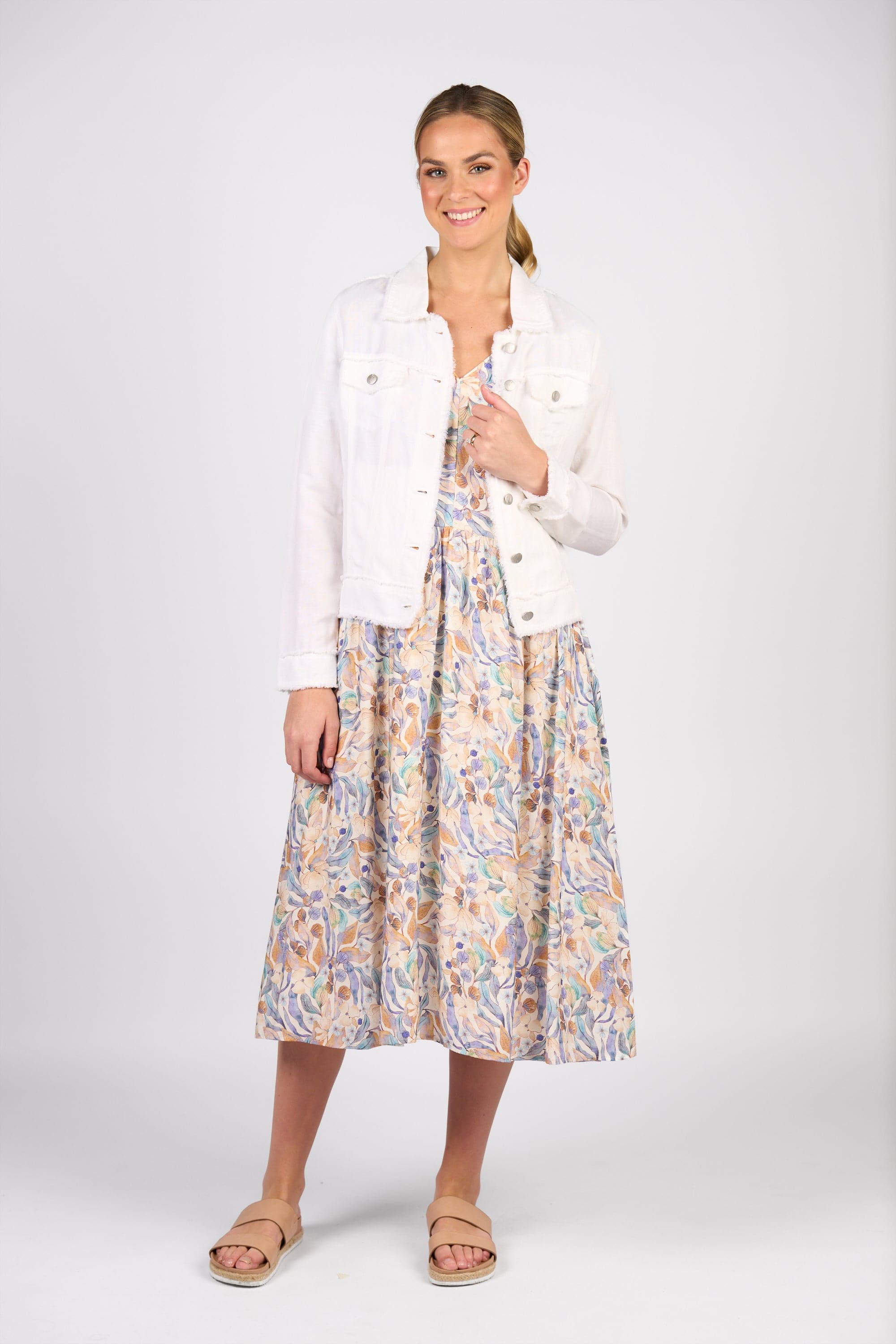 A woman smiles in the Vassalli Aura Jacket - White, styled over a pastel patterned midi dress and beige sandals. Her hair is tied back as she poses against a plain light background.