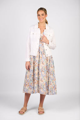 A woman smiles in the Vassalli Aura Jacket - White, styled over a pastel patterned midi dress and beige sandals. Her hair is tied back as she poses against a plain light background.