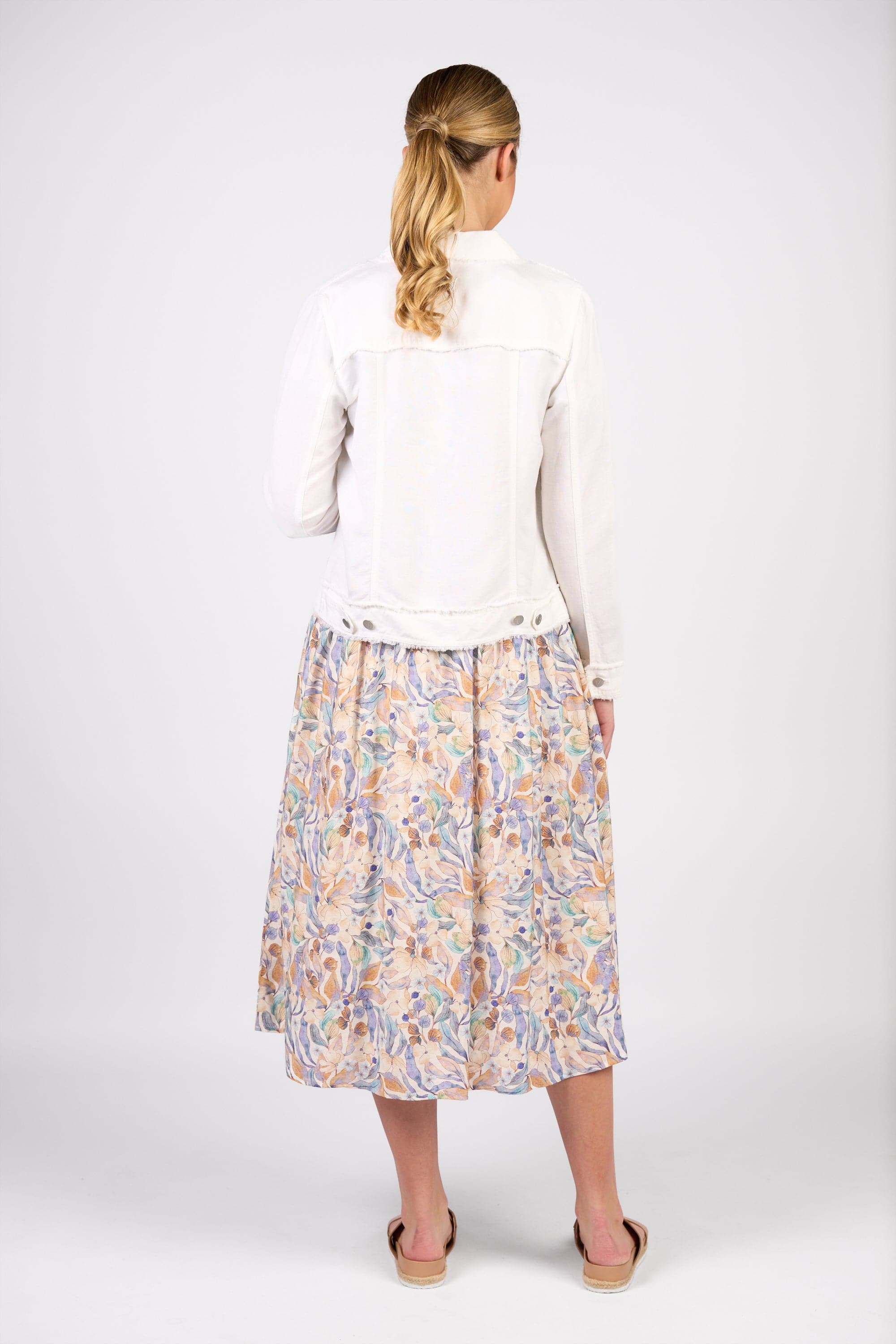 A woman with blonde hair in a low ponytail faces away, wearing the Vassalli Aura Jacket in white and a pastel floral midi skirt with sandals, standing against a plain white background.