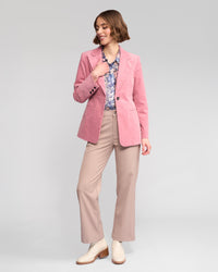 A woman smiles to her left, hand on hip, wearing the Vassalli Gallery Blazer in dusky pink against a plain background.