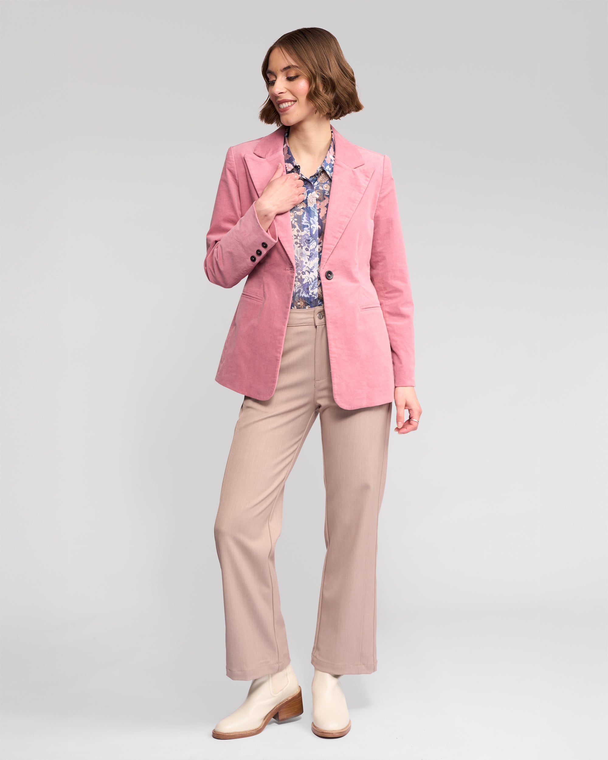 A woman smiles to her left, hand on hip, wearing the Vassalli Gallery Blazer in dusky pink against a plain background.