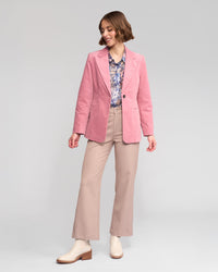A woman wears the Vassalli Gallery Blazer in dusky pink, paired with a floral blouse and beige pinwale cord pants. She smiles, looking down to her left, posed against a plain light background with one foot forward.