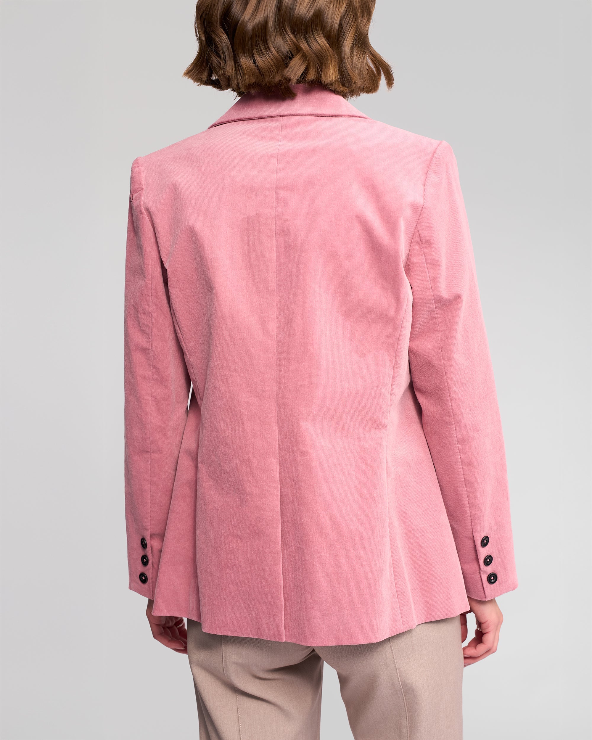 A woman with short brown hair is seen from behind wearing the Vassalli Gallery Blazer in dusky pink with black sleeve buttons and beige pants, set against a plain light gray background.