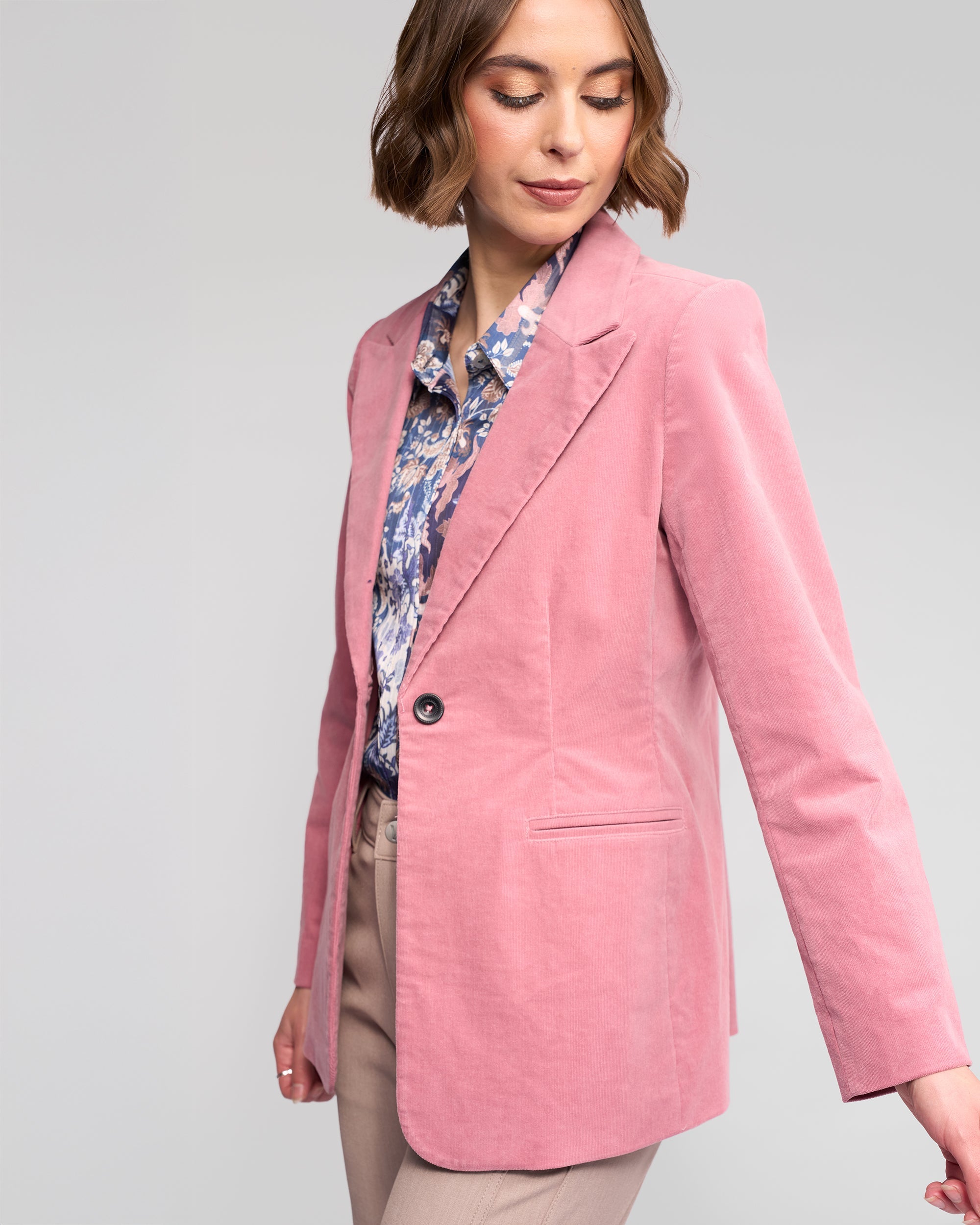 A woman wears the Vassalli Gallery Blazer in dusky pink, paired with a blue floral blouse and beige pinwale cord pants, standing against a plain light gray background and looking down with a slight smile.