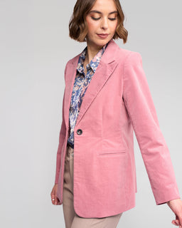 A woman wears the Vassalli Gallery Blazer in dusky pink, paired with a blue floral blouse and beige pinwale cord pants, standing against a plain light gray background and looking down with a slight smile.