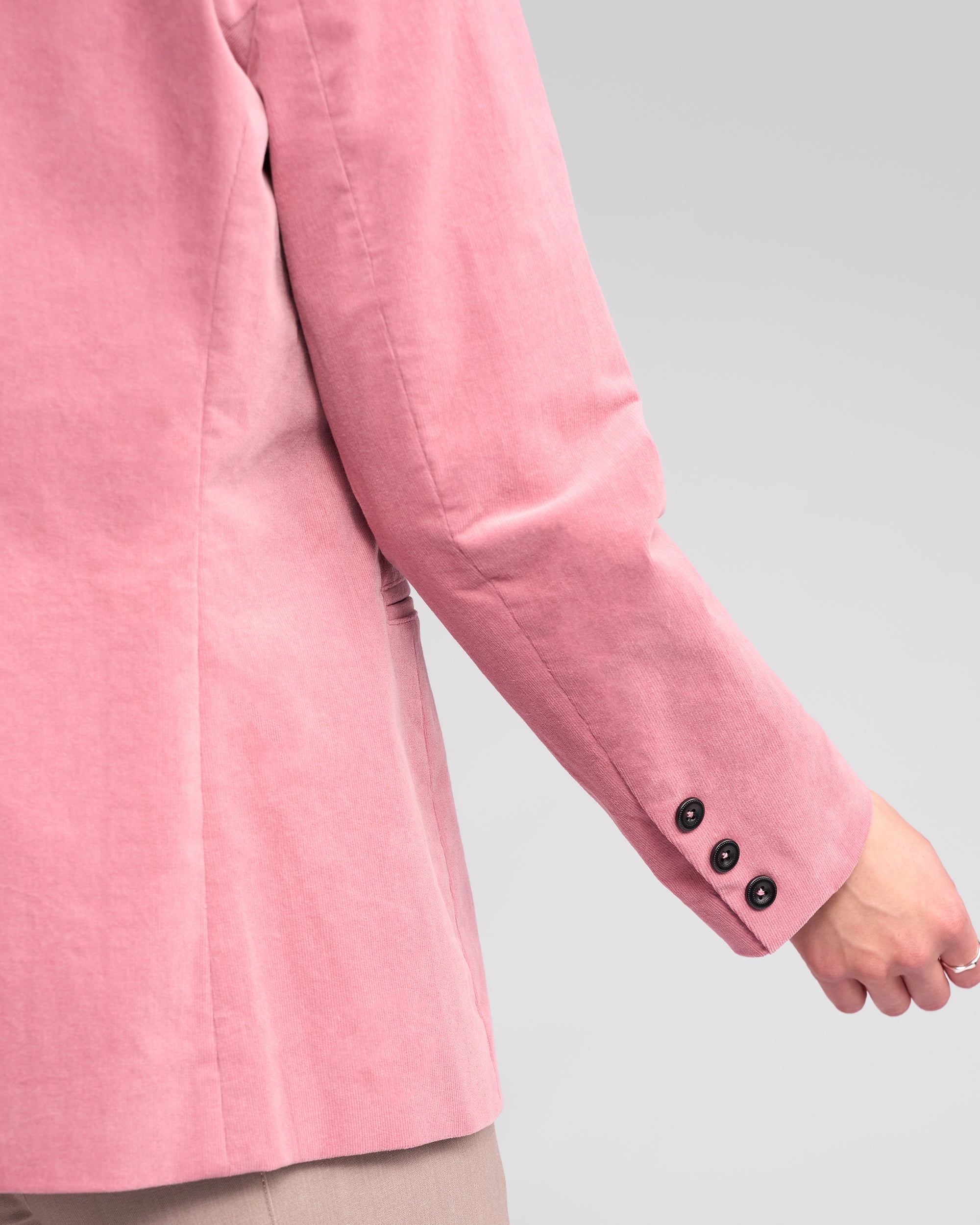 A person wears the Vassalli Gallery Blazer in dusky pink, featuring black sleeve buttons and a soft pinwale cord texture, shown from the back and side with a slightly bent arm, set against a plain light gray background.