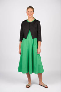 A woman stands smiling, wearing a bright green midi dress with a Vassalli Cropped Jacket in black and flat, patterned sandals against a plain white background.