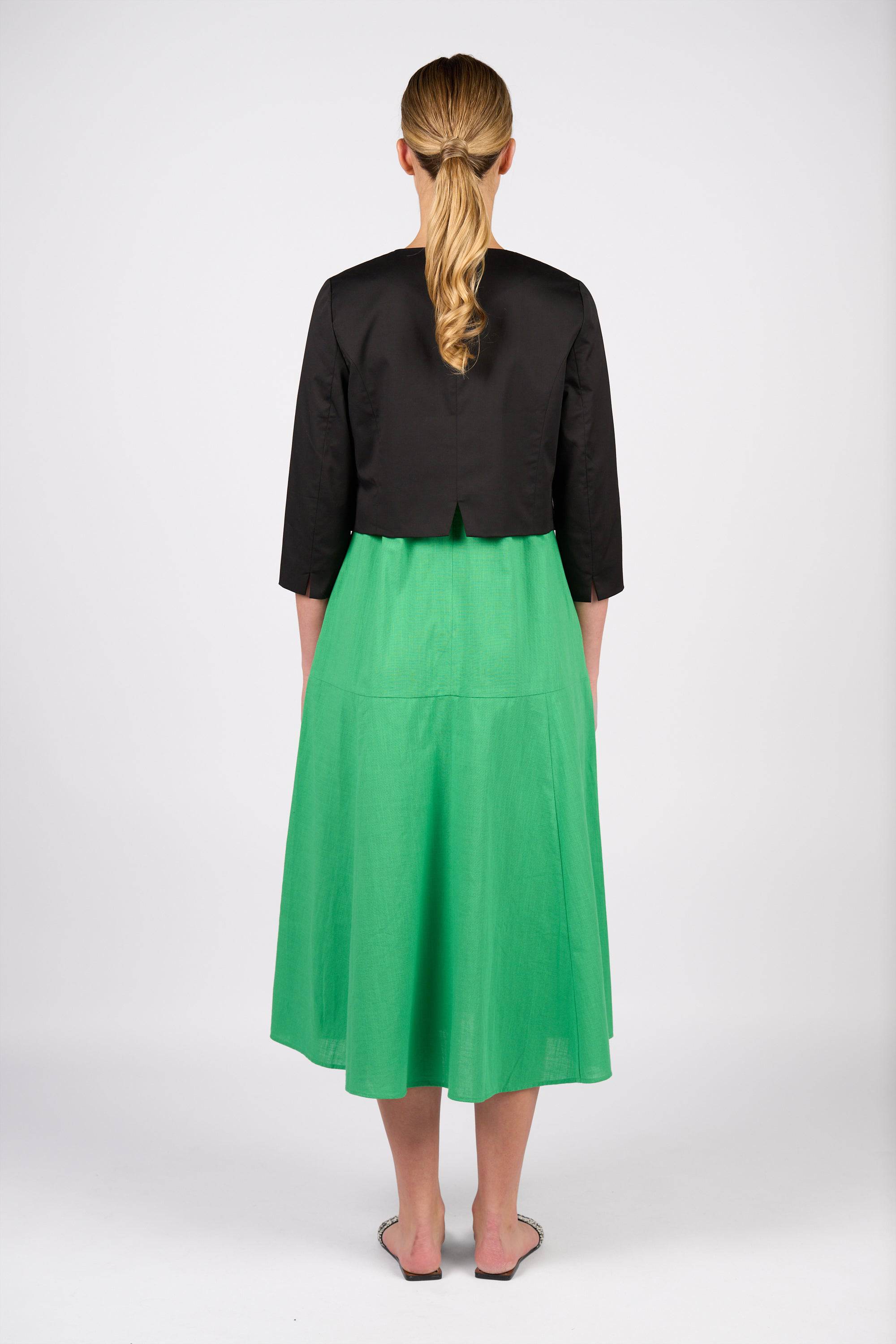 A woman with blonde hair in a low ponytail, seen from behind against a white background, wears a Vassalli Cropped Jacket - Black with 3/4 sleeves, a bright green midi skirt, and pointed flats.