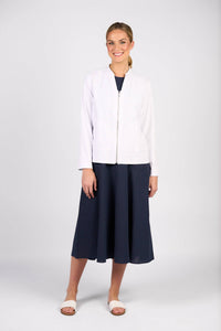 A woman smiles against a plain background, wearing the Vassalli Zip Up Jacket in white over a knee-length navy dress and white sandals.