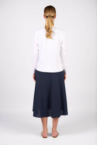 A woman with blonde hair in a ponytail faces away, wearing the Vassalli Zip Up Jacket in white, a dark blue midi skirt, and white slip-on shoes against a plain white background.