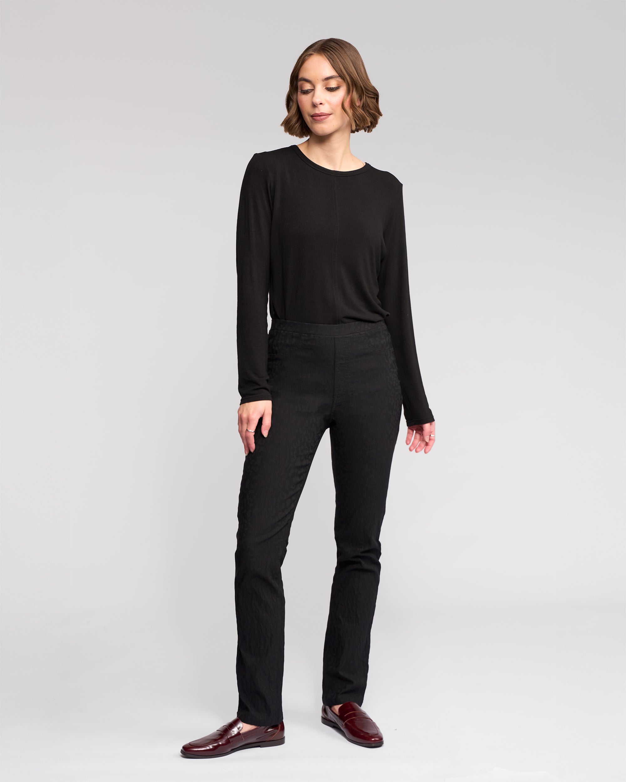 A woman with short brown hair stands against a plain background, wearing Vassalli's Bay Pull On - Black pants and a long-sleeve black top, paired with dark red loafers. She looks slightly to the side with a relaxed expression.