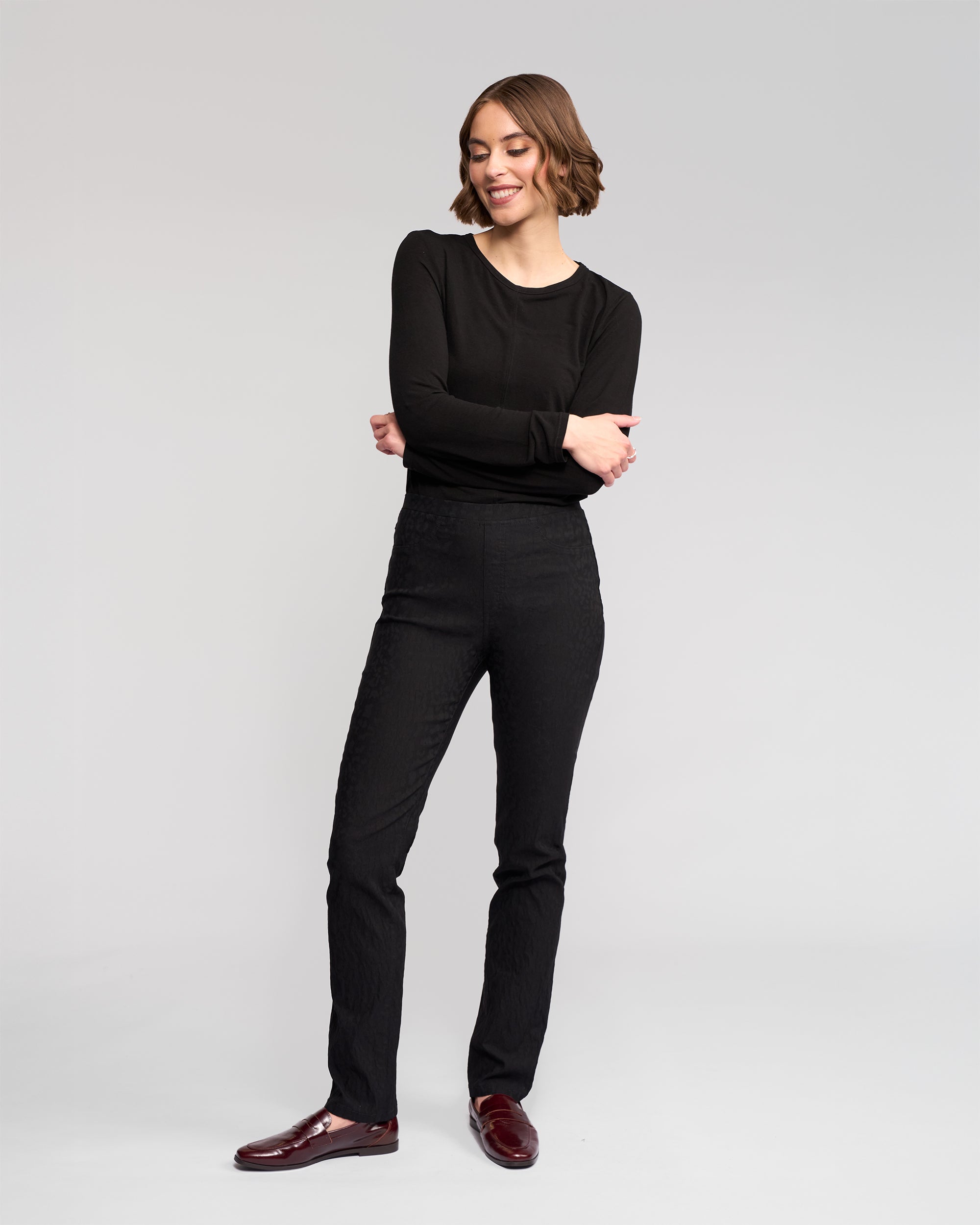 A woman with short brown hair, smiling with arms crossed, wears a black long-sleeve top and Vassalli Bay Pull On - Black pants with burgundy shoes, standing against a plain light gray background.