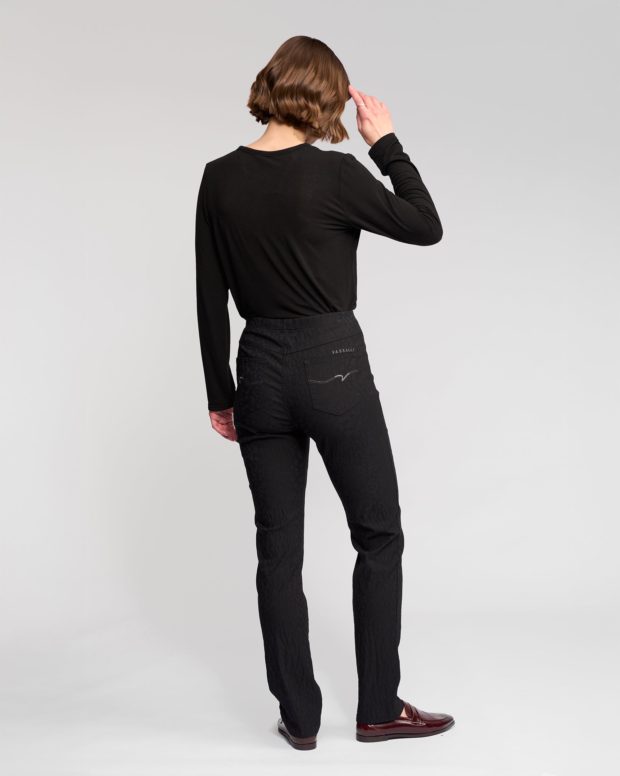 A person with short brown hair wears Vassalli's Bay Pull On - Black pants, a black long-sleeve shirt, and brown shoes, facing away and slightly to the side against a plain light gray background.