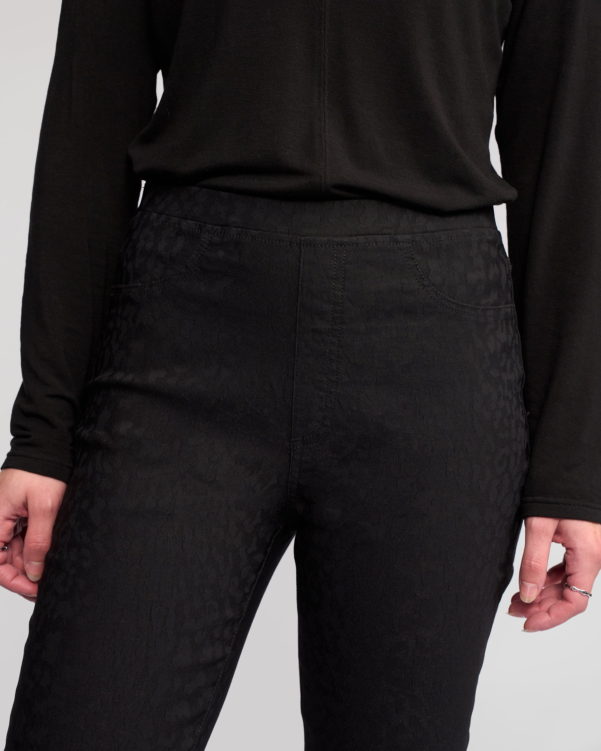 A person models the Vassalli Bay Pull On - Black, a long-sleeved top and high-waisted slim pants with subtle animal texture, shown from shoulders to mid-thigh. Hands rest at their sides; one hand wears rings.