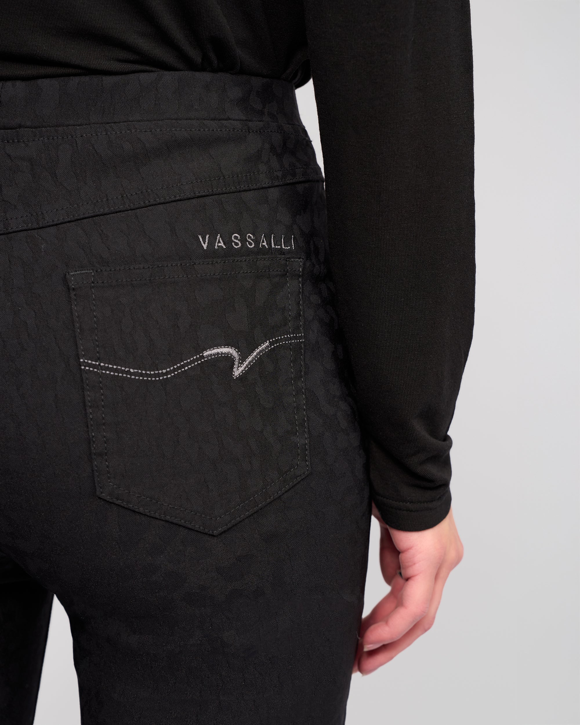 A close-up of a person wearing Vassalli Bay Pull On - Black pants, featuring animal-textured fabric, a pull-on style, and a back pocket with VASSALLI embroidery and white stitching.