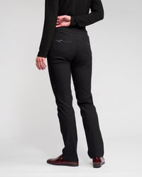 A person stands facing away against a plain, light background, wearing Vassalli's Bay Pull On - Black slim pants with a black long-sleeve top and burgundy loafers. One hand is resting behind their back.