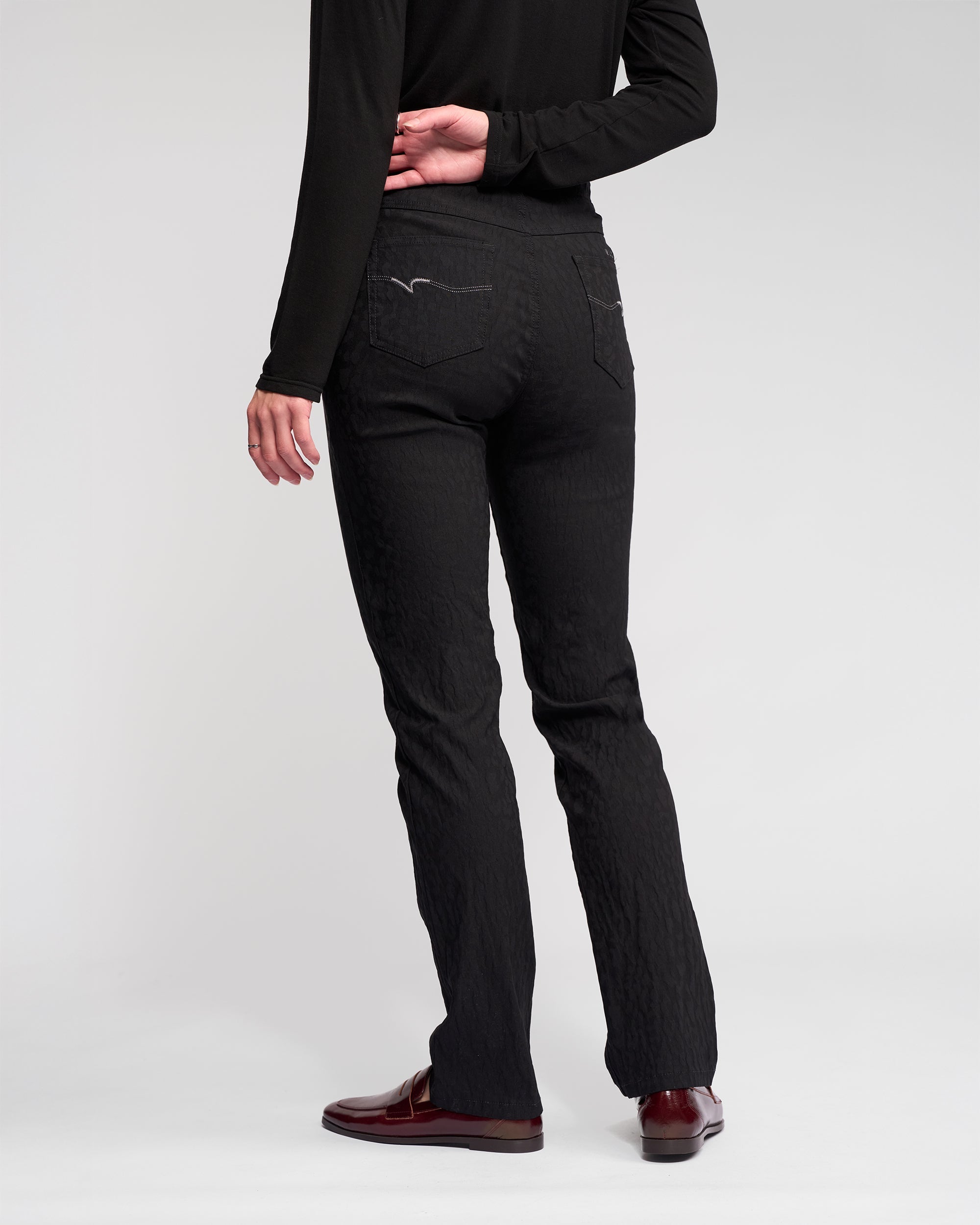 A person stands facing away against a plain, light background, wearing Vassalli's Bay Pull On - Black slim pants with a black long-sleeve top and burgundy loafers. One hand is resting behind their back.