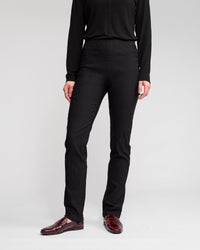 A person stands against a plain background, wearing the Vassalli Bay Pull On - Black long-sleeve top, animal-textured slim pants, and burgundy loafers. Their face is not visible in the image.