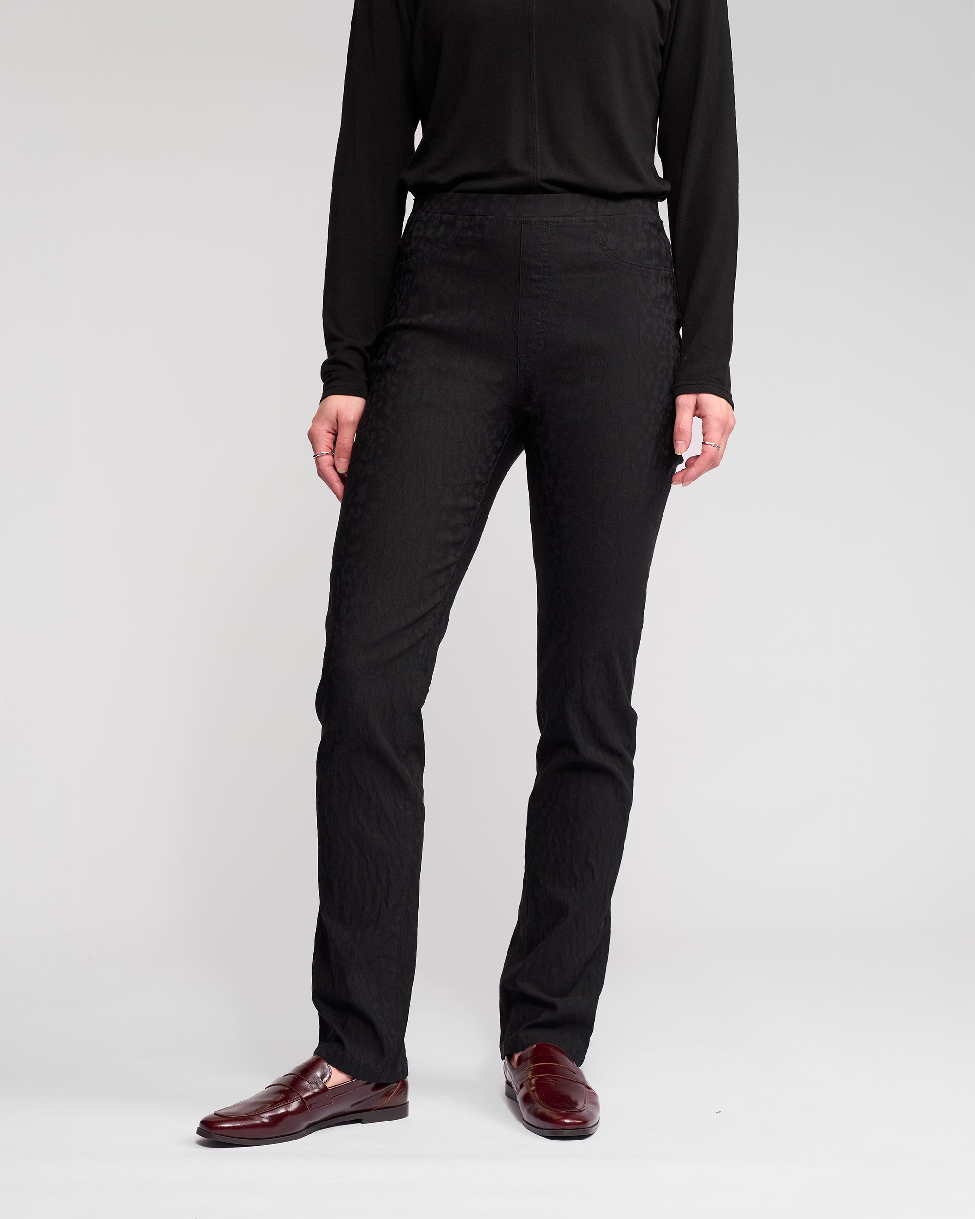 A person stands against a plain background, wearing the Vassalli Bay Pull On - Black long-sleeve top, animal-textured slim pants, and burgundy loafers. Their face is not visible in the image.