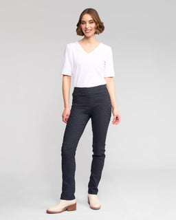 A woman with short brown hair, dressed in a white V-neck T-shirt, light ankle boots, and Vassalli Bay Pull On - Navy slim animal-textured pants with an elastic waist, smiles against a plain light gray background.