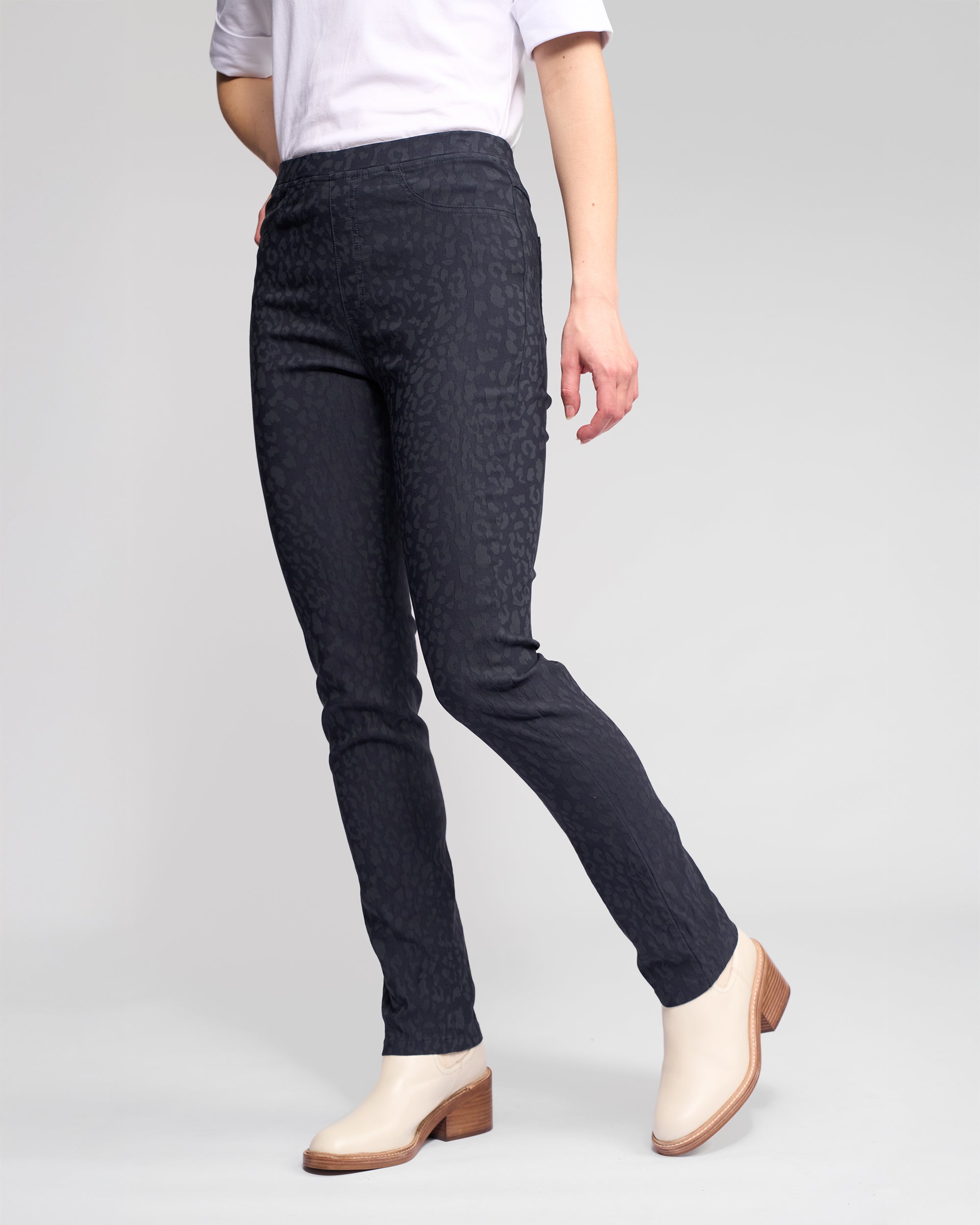 A person wearing Vassalli Bay Pull On - Navy slim pants with an elastic waistband, a white top with sleeves rolled up, and cream ankle boots stands against a light gray background. The person's head is not visible.