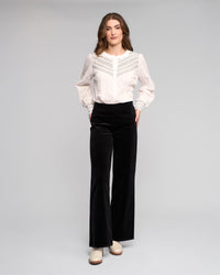 A woman stands against a light background wearing a white lace-detailed blouse and Vassalli Loft Cord Pull On pants in black, featuring a wide-leg design and elastic waistband. She faces forward with her hands in the pockets, paired with light heeled boots.