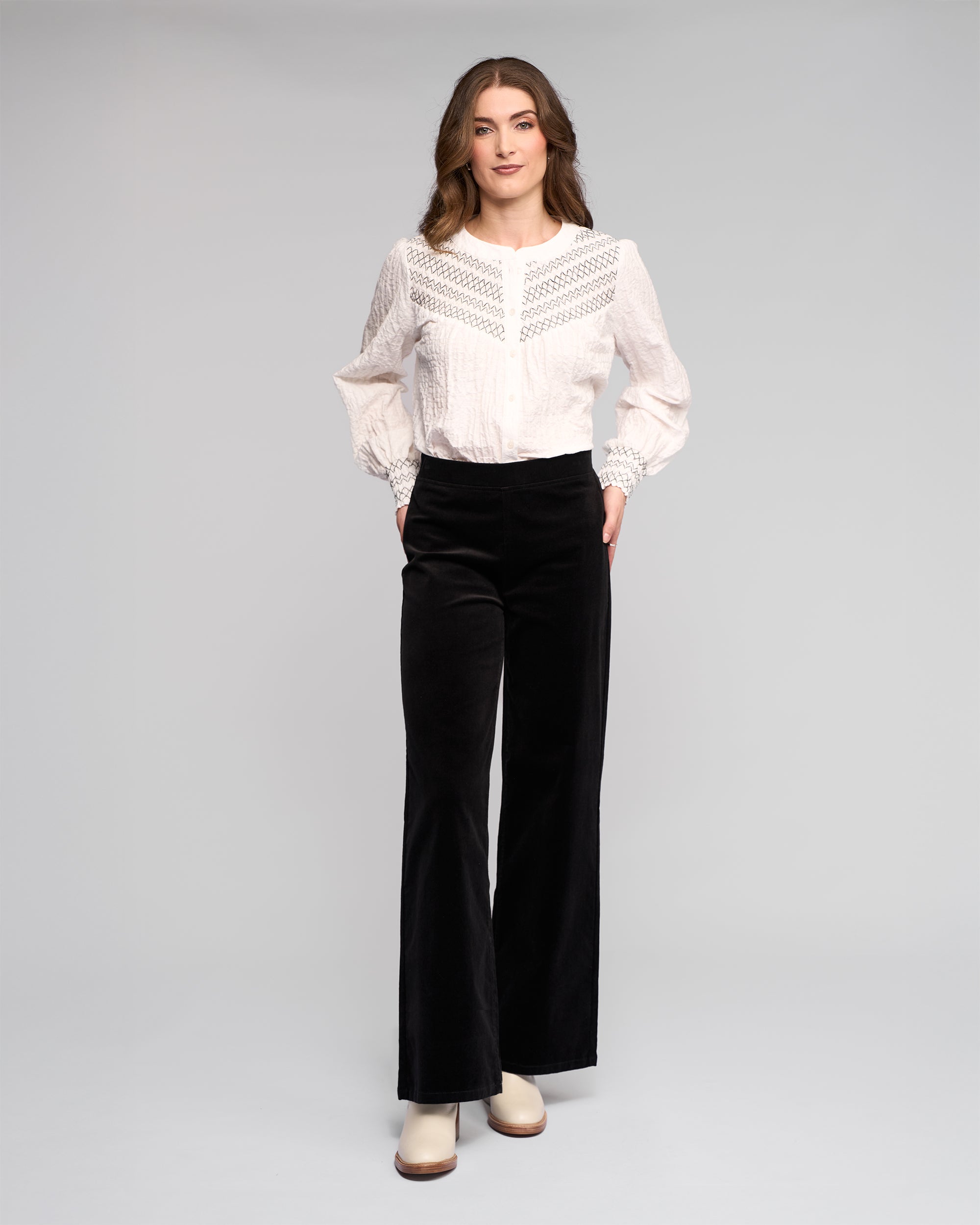 A woman stands against a light background wearing a white lace-detailed blouse and Vassalli Loft Cord Pull On pants in black, featuring a wide-leg design and elastic waistband. She faces forward with her hands in the pockets, paired with light heeled boots.