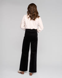 A woman with wavy brown hair faces away, wearing a textured white long-sleeve blouse and Vassalli Loft Cord Pull On - Black wide-leg pants with an elasticated waistband, plus beige heeled shoes, against a plain light gray background.