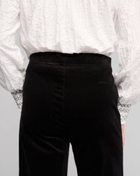 Model wears a textured white long-sleeve shirt with patterned cuffs and Vassalli Loft Cord Pull On - Black pinwale cord wide-leg pants, shown from the back. Subtle text detail is visible near the waistband on the right side of the pants.