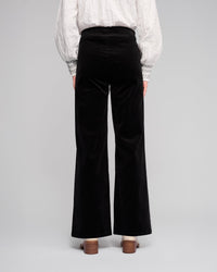A person stands facing away, wearing Vassalli's Loft Cord Pull On - Black pants with an elasticated waistband, a white textured blouse with patterned cuffs, and brown heeled shoes against a plain light background.