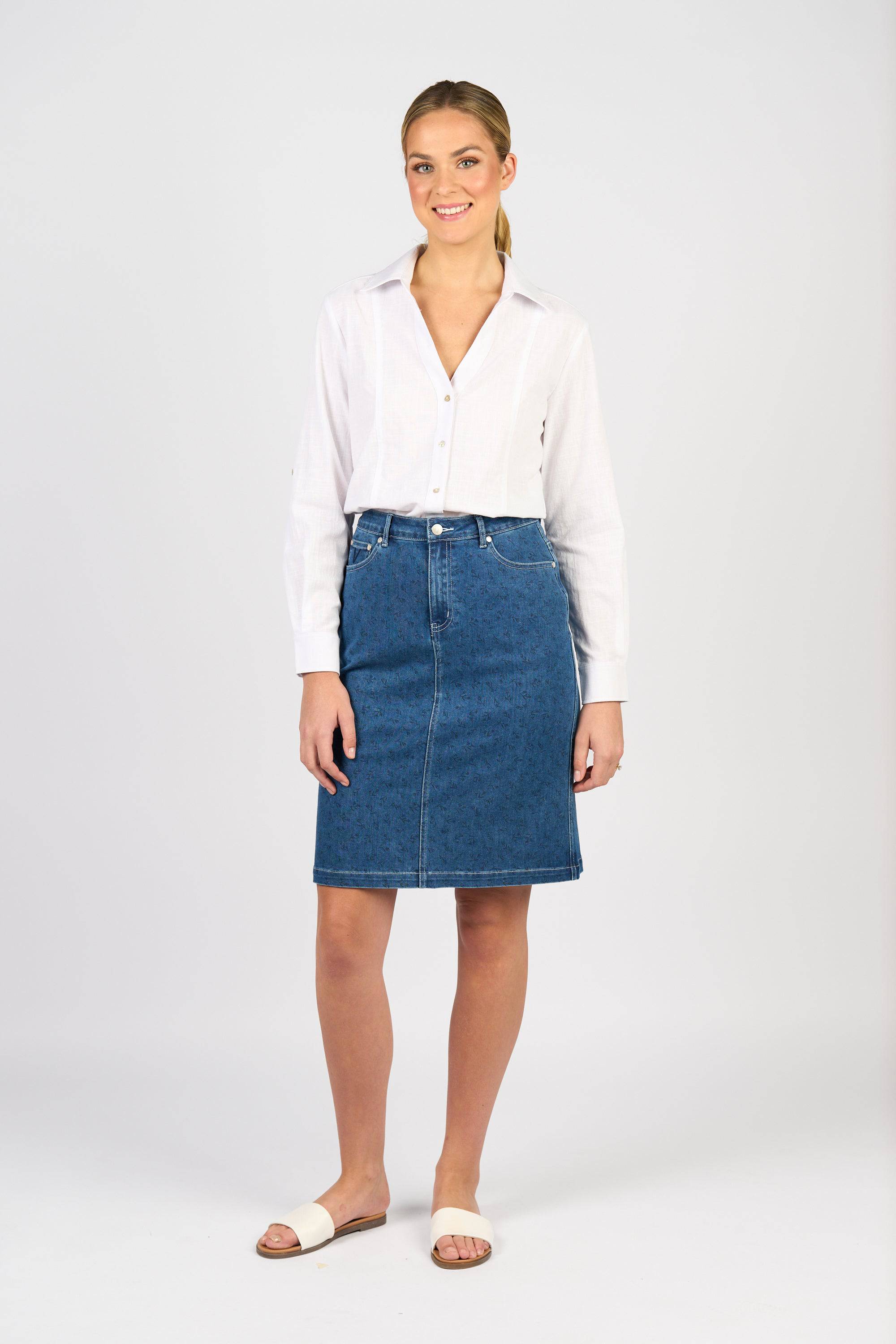 A person stands against a plain background, smiling at the camera and wearing a white long-sleeve button-up shirt, white slide sandals, and the Vassalli Poppy Skirt in Kansas Denim.