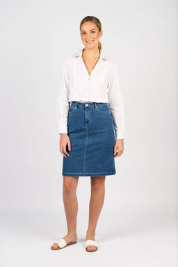 A person stands against a plain background, smiling at the camera and wearing a white long-sleeve button-up shirt, white slide sandals, and the Vassalli Poppy Skirt in Kansas Denim.
