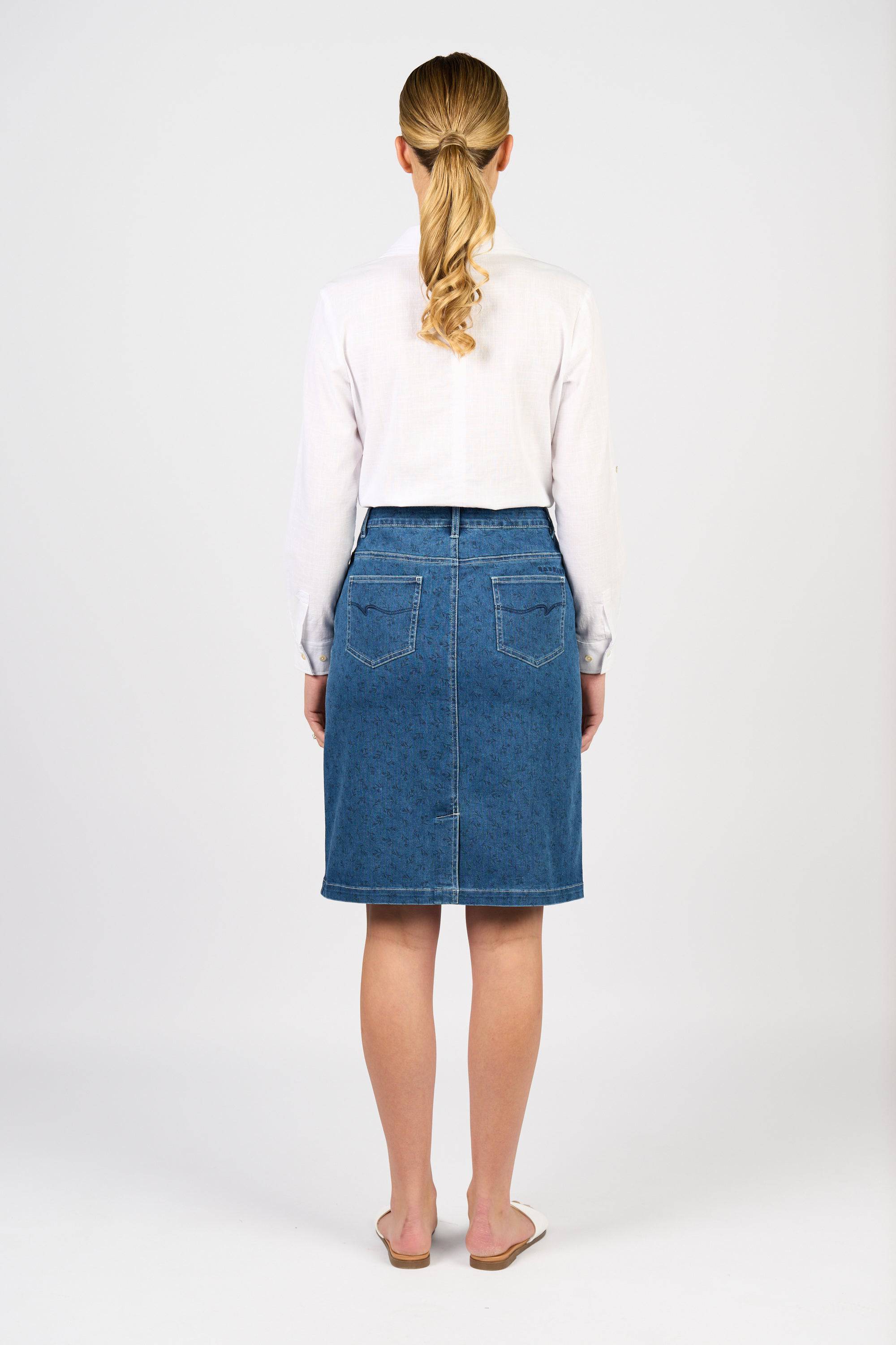 A woman with blonde hair in a ponytail stands facing away, wearing a white long-sleeve shirt, the Vassalli Poppy Skirt - Kansas Denim, and white flat shoes against a plain white background.