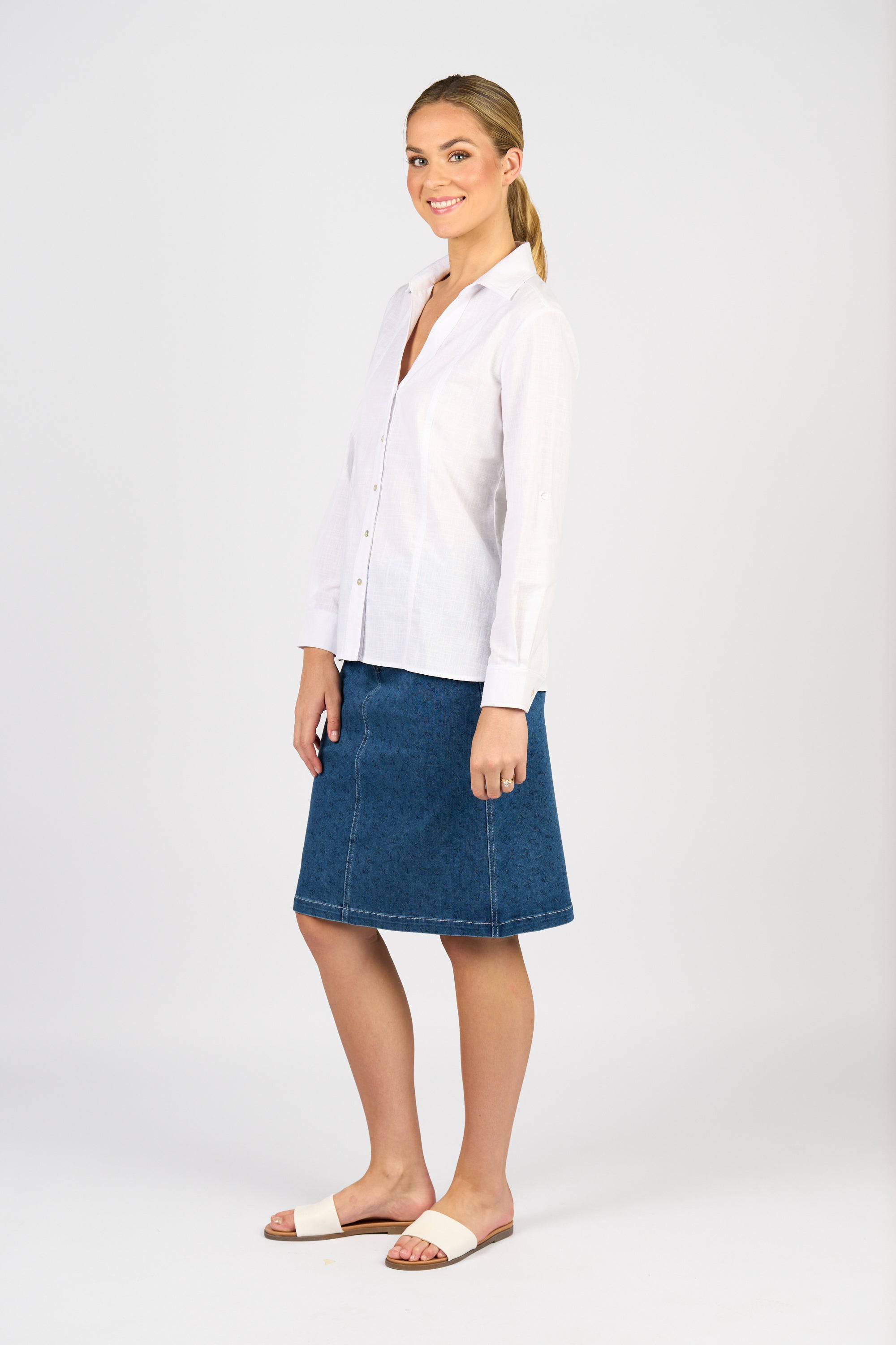 A woman smiles in a white button-up shirt, Vassalli Poppy Skirt - Kansas Denim, and white slide sandals, posed at an angle against a plain white background.