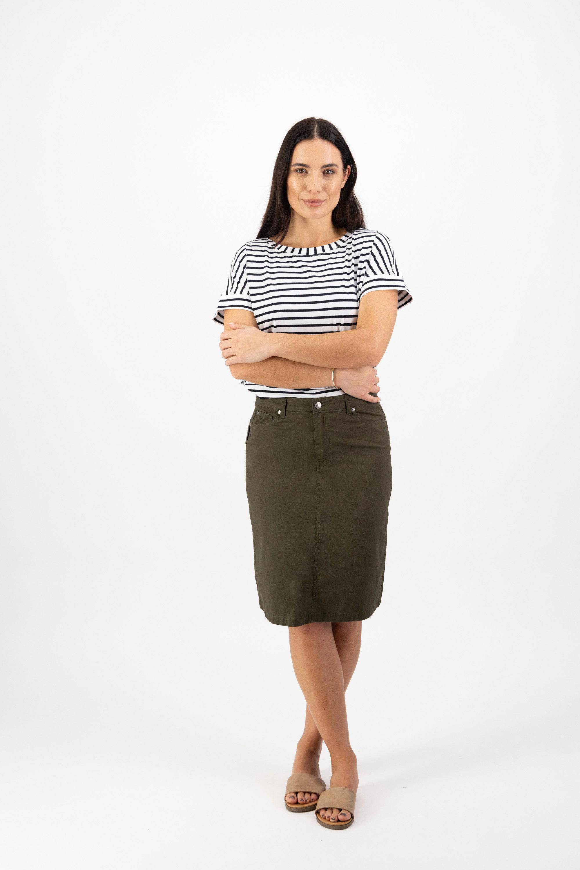 A woman with long dark hair stands against a white background, wearing a black and white striped t-shirt, the Vassalli Poppy Poplin Skirt in Khaki, and beige sandals. Her arms are crossed and she looks at the camera.