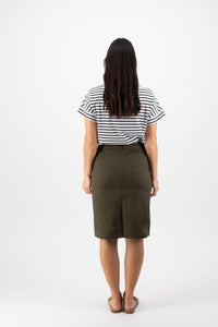 A woman, seen from behind, wears a black and white striped shirt, brown sandals, and the Vassalli Poppy Poplin Skirt in khaki, standing against a plain white background.