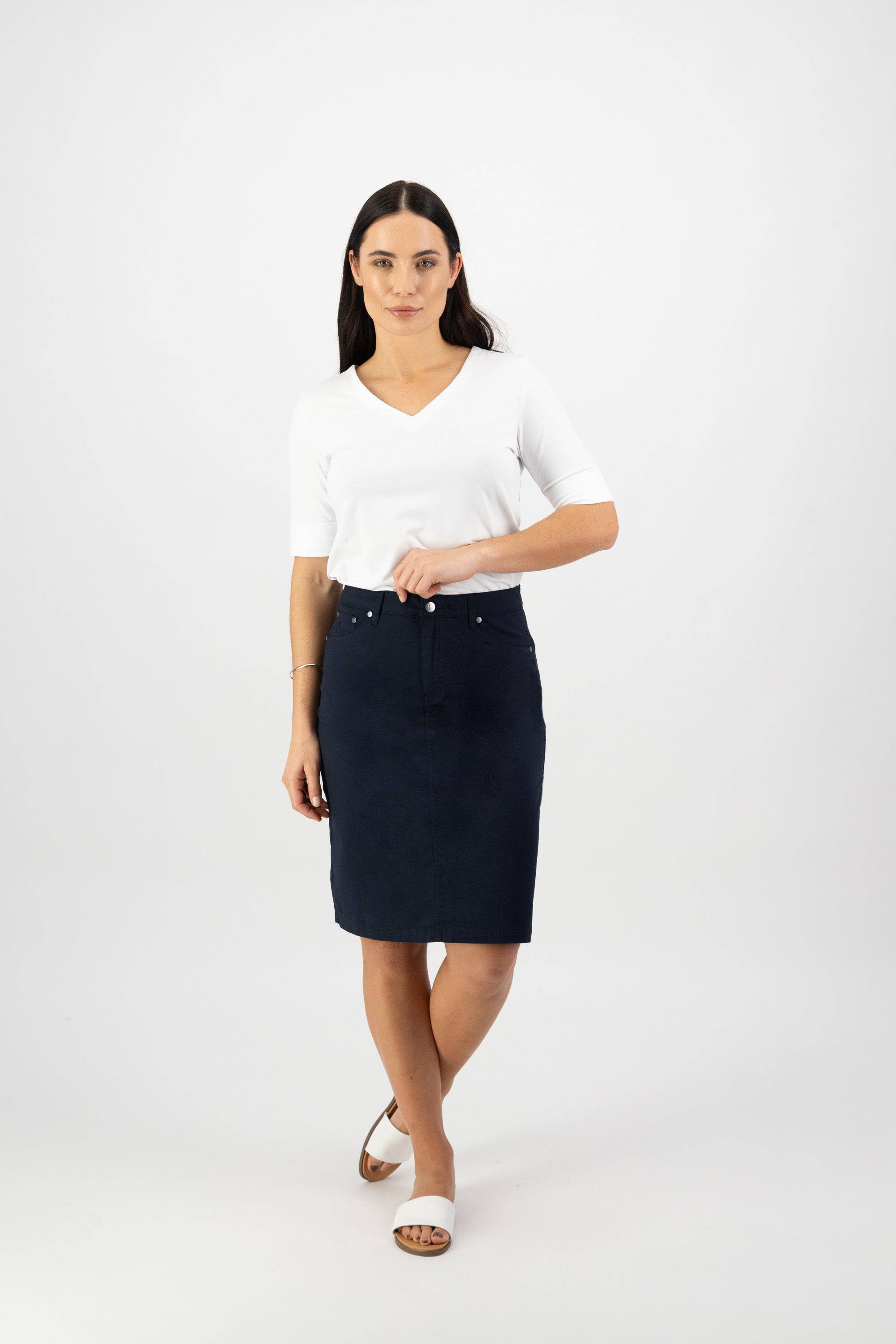 A woman with long dark hair wears a white V-neck top, white sandals, and the Vassalli Poppy Poplin Skirt in navy. She stands against a plain white background, facing forward with one arm slightly bent.