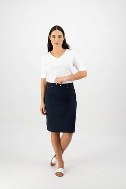 A woman with long dark hair wears a white V-neck top, white sandals, and the Vassalli Poppy Poplin Skirt in navy. She stands against a plain white background, facing forward with one arm slightly bent.