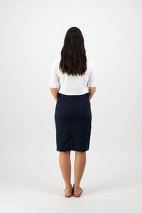 A woman with long dark hair faces away, wearing a white top, sandals, and the Vassalli Poppy Poplin Skirt in Navy, set against a plain white background.
